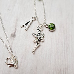 Op de afbeelding: Zilverkleurige ketting met een feeënbedel, een groene edelsteen en de letter "A". De fee heeft gedetailleerde vleugels en een delicate pose. De ketting wordt gepresenteerd op een licht oppervlak met groene bladeren op de achtergrond.