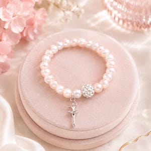 May include: A bracelet made of pink pearls, a crystal bead, and a silver ballerina charm. The bracelet is displayed on a pink velvet surface, with pink flowers in the background. A charming piece of jewellery.