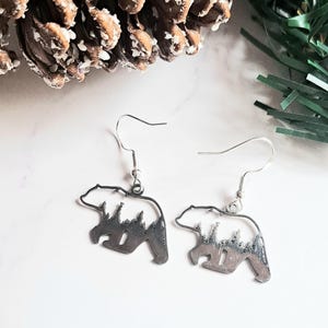 以下が含まれることがあります： 銀色のクマの形をしたイヤリングのペア。各イヤリングは、内部に切り抜かれた森のシーンがあるクマのシルエットが特徴です。イヤリングは、銀色のフィッシュフックのイヤワイヤーから吊り下げられています。イヤリングは白い大理石の表面に表示されています。