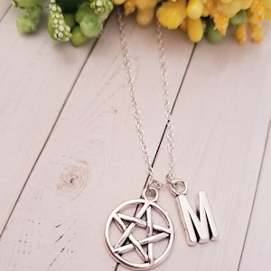 Puede incluir: Un collar de cadena de plata con dos colgantes: un colgante de pentagrama y un colgante de letra "M". El collar está sobre una superficie de madera blanca.