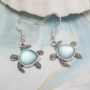 Puede incluir: Un par de pendientes de tortuga plateados con detalles de vidrio iridiscente azul claro. Los pendientes están sobre una superficie de mármol blanco y azul.