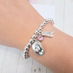 May include: Silver beaded bracelet with cowboy-themed charms. The bracelet features a cowboy hat, a cowboy boot, and a pink crystal charm. The bracelet is worn on a wrist.