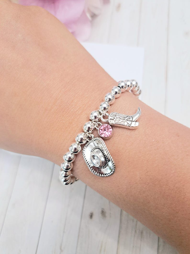 May include: Silver beaded bracelet with cowboy-themed charms. The bracelet features a cowboy hat, a cowboy boot, and a pink crystal charm. The bracelet is worn on a wrist.