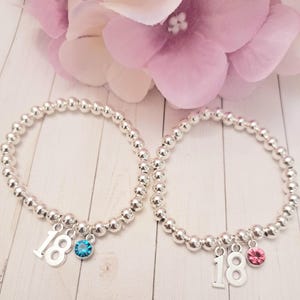 Peut inclure: Deux bracelets de perles argentées avec des breloques. Chaque bracelet comporte un chiffre 18 en argent et une breloque en pierre précieuse. Une pierre est bleue, l'autre rose. Les bracelets sont devant une fleur rose.