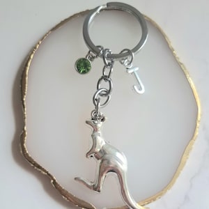 May include: Silver-toned keychain featuring a kangaroo charm, a green gemstone, and a letter "J" pendant. The keychain is attached to a silver ring. The kangaroo charm is detailed and stands out against the white background.