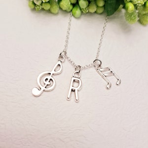 Puede incluir: Un collar de plata con tres colgantes: una clave de sol, una nota musical y la letra "R".
