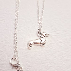 May include: A silver necklace with a dachshund charm. The necklace is on a white surface. The charm is detailed and has a 925 stamp.