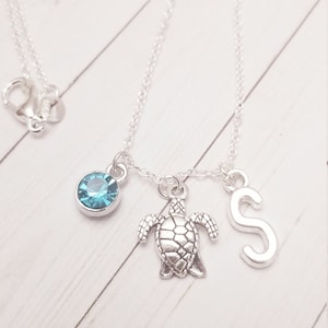 Könnte beinhalten: Silberkette mit drei Anhängern: einem runden blauen Edelstein, einer Schildkröte und dem Buchstaben "S". Die Kette wird auf einer weißen Holzoberfläche präsentiert.