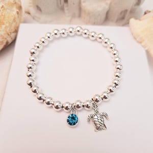 Könnte beinhalten: Silbernes Perlenarmband mit einem blauen Edelstein-Anhänger und einem Schildkröten-Anhänger. Das Armband wird auf einer weißen Oberfläche mit Muscheln und einer Sandburg im Hintergrund präsentiert. Das Armband besteht aus kleinen, runden, silbernen Perlen.