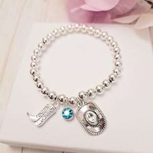 May include: Silver beaded bracelet with cowboy-themed charms. The charms include a cowboy boot, a turquoise blue crystal, and a cowboy hat. The bracelet is displayed on a white surface with a pink flower in the background.