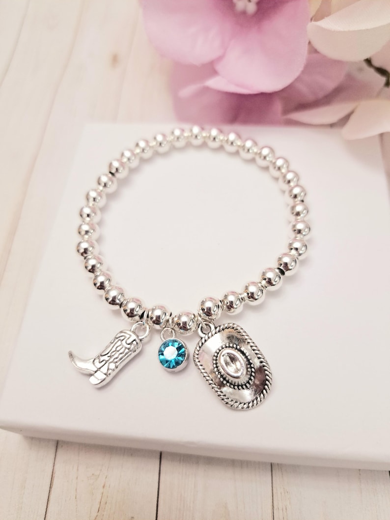 May include: Silver beaded bracelet with cowboy-themed charms. The charms include a cowboy boot, a turquoise blue crystal, and a cowboy hat. The bracelet is displayed on a white surface with a pink flower in the background.