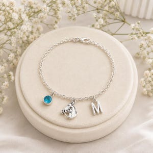 May include: A silver-toned chain bracelet featuring three dangling charms: a round blue crystal, a detailed horse head profile, and the capital letter M. The bracelet is constructed with a delicate link chain and a secure lobster claw clasp.