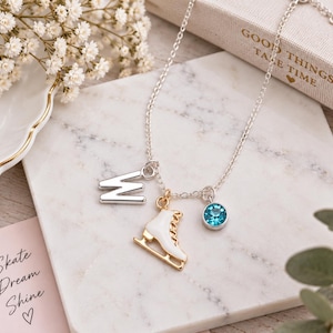 May include: A silver-toned chain necklace featuring three charms: a silver letter W, a white and gold-toned ice skate, and a round, bright blue crystal. The necklace is displayed on a white marble surface near a book with the text Good Things Take Time.