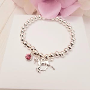 May include: Silver beaded bracelet featuring a pink gemstone and a silver horse charm. The bracelet is composed of small, round, silver beads. The charm is a detailed horse. The gemstone is a round, pink jewel.