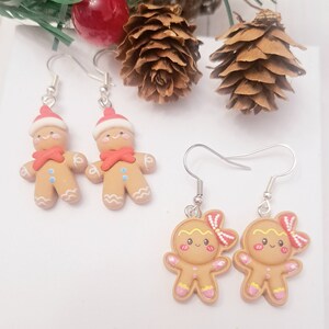 May include: Two pairs of festive gingerbread man earrings. One pair features gingerbread men wearing Santa hats and red scarves. The other pair features gingerbread women with pink bows. The earrings are on silver hooks, perfect for holiday wear.