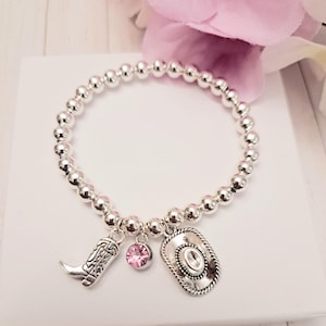 May include: Silver beaded bracelet with cowboy-themed charms. The bracelet features a cowboy boot, a pink gemstone, and a cowboy hat charm. The bracelet is displayed on a white surface with a pink flower in the background.