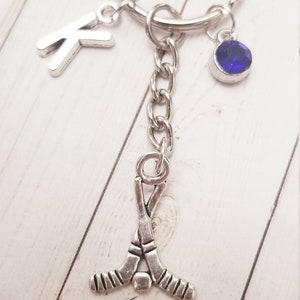 May include: Silver-toned hockey keychain with a letter "K" charm, a blue gemstone, and crossed hockey sticks. The keychain is attached to a silver key ring. The background is a light wood grain.
