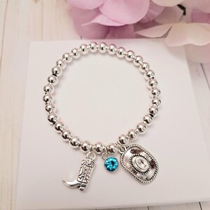 May include: Silver beaded bracelet with cowboy-themed charms. The bracelet features a cowboy boot, a cowboy hat, and a blue gemstone charm. The bracelet is displayed on a white surface with a pink flower in the background.