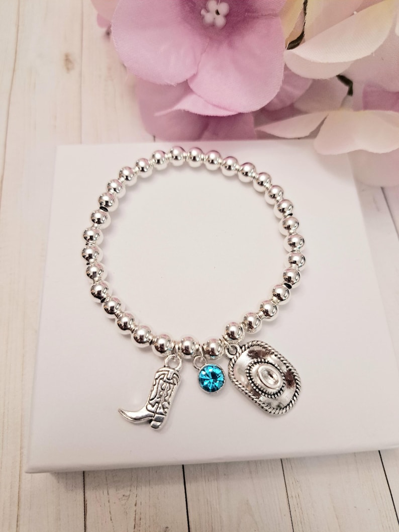 May include: Silver beaded bracelet with cowboy-themed charms. The bracelet features a cowboy boot, a cowboy hat, and a blue gemstone charm. The bracelet is displayed on a white surface with a pink flower in the background.