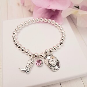 May include: Silver beaded bracelet with a cowboy boot charm, a pink gemstone charm, and a cowboy hat charm. The bracelet is displayed on a white surface with pink flowers in the background.
