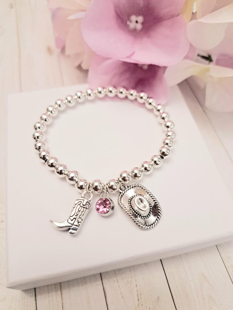 May include: Silver beaded bracelet with a cowboy boot charm, a pink gemstone charm, and a cowboy hat charm. The bracelet is displayed on a white surface with pink flowers in the background.