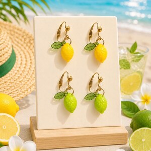 May include: Four pairs of lemon-shaped earrings on display. Two pairs are yellow, and two are green, each with a small green leaf and gold-colored hardware. The earrings are presented on a white stand with a wooden base, with a beach scene in the background.