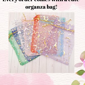 May include: A collection of colorful organza bags with gold drawstrings, decorated with gold stars and crescent moons. The bags are in shades of green, blue, purple, red, and white. Text reads: Every order comes with a cute organza bag!
