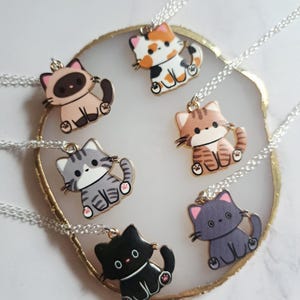 May include: A collection of seven cat-shaped pendant necklaces. Each pendant features a different color scheme, including black, gray, brown, and calico. The necklaces have silver-colored chains and are arranged on a gold-rimmed, white and gold surface.