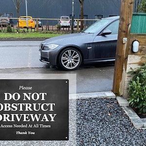 May include: A black and white sign with the text "Please Do Not Obstruct Driveway Access Needed At All Times Thank You" on a black background. A gray car is driving past the sign.
