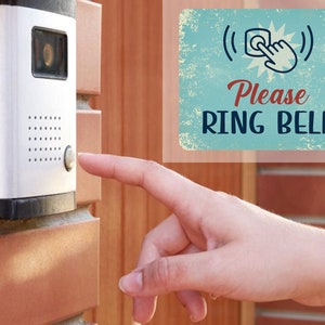 May include: A silver doorbell with a black button and a small camera lens. A hand is pressing the button. A vintage-style sign reads "Please Ring Bell" with a stylized bell and hand icon.