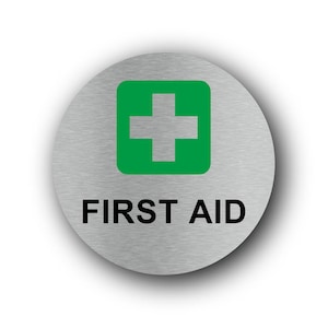 May include: A circular brushed metal sign featuring a green square with a white cross symbol centred at the top. Below the green square, the text FIRST AID is printed in bold, black, sans-serif capital letters on the silver surface.