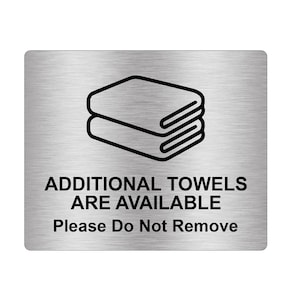 May include: A silver metal sign with a black outline of two towels stacked on top of each other. The sign reads "ADDITIONAL TOWELS ARE AVAILABLE Please Do Not Remove".
