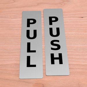 Peut inclure: Deux panneaux rectangulaires argentés avec des lettres noires. Le panneau de gauche indique "PULL" et le panneau de droite indique "PUSH".