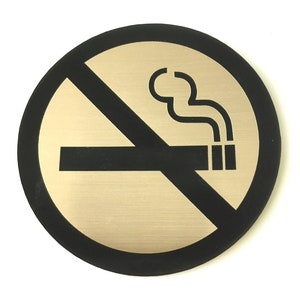 Puede incluir: Un cartel redondo de color negro y dorado con la señal de prohibido fumar, con una línea diagonal que atraviesa un cigarrillo del que sale humo.
