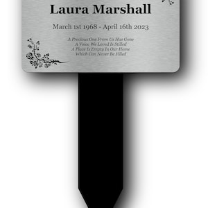 May include: A rectangular silver-toned metal memorial plaque with a black stake. The plaque features the text In Loving Memory of Laura Marshall, March 1st 1968 - April 16th 2023, and a poem, decorated with black rose vine illustrations at the corners.