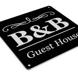 Puede incluir: Un letrero cuadrado negro con letras blancas que dice "B&B Guest House". El letrero tiene un adorno decorativo sobre el "B&B".