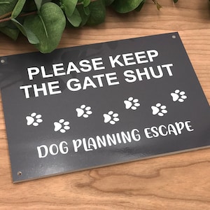 May include: Black sign with white text that reads "Please keep the gate shut" with paw prints and the text "Dog planning escape".
