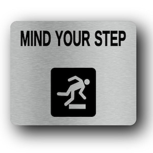Puede incluir: Un letrero rectangular de metal cepillado con esquinas redondeadas. Las palabras "MIND YOUR STEP" están impresas en negrita, en letras negras en la parte superior. Debajo, un cuadrado negro contiene un gráfico blanco de una persona bajando.