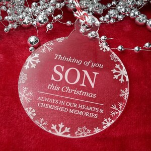 May include: A clear round Christmas bauble with a white snowflake border. The bauble has the text "Thinking of you SON this Christmas ALWAYS IN OUR HEARTS & CHERISHED MEMORIES" printed on it.