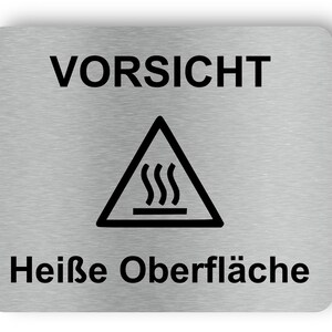 May include: A rectangular, brushed metal sign with rounded corners. The word "VORSICHT" is at the top, and "Heiße Oberfläche" is at the bottom in black text. A black triangle with a steam symbol inside is in the center.
