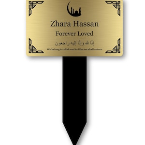 May include: A gold-colored memorial stake with black accents. The stake features the name "Zhara Hassan" and the words "Forever Loved" in black text. It also includes Arabic script and the English phrase "We belong to Allah and to Him we shall return."