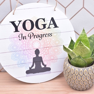 May include: A white round wooden sign with the text "YOGA In Progress" and a black silhouette of a person in a yoga pose. The sign has a rainbow mandala design behind the person.