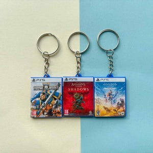 Op de afbeelding: Drie PS5 game case sleutelhangers. De sleutelhangers tonen de speltitels: Space Marine, Assassin's Creed Shadows en Horizon. Elke sleutelhanger heeft een blauwe rand en een zilveren sleutelring.