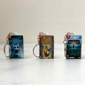 May include: Three miniature video game case keychains. The keychains feature the game titles "Hollow Knight", "It Takes Two", and "Bioshock". Each keychain has a silver keyring attached. The background is a light, neutral color.