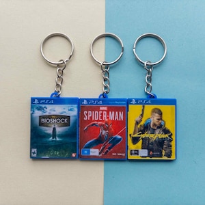 May include: Three PS4 game case keyrings. The keyrings feature the game titles "Bioshock", "Spider-Man", and "Cyberpunk". Each keyring has a blue plastic case with a silver ring attached. The background is a two-tone blue.