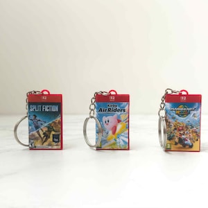 May include: Three red keychains featuring video game box art. The keychains have silver key rings and chains. The game titles are "Split Fiction", "Kirby Air Riders", and "Mario Kart 8 Deluxe".