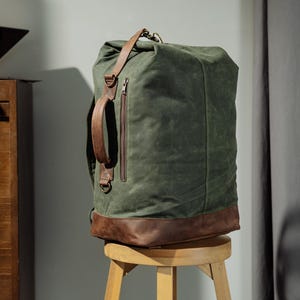 Pode incluir: Uma mochila de lona verde-oliva escura com base de couro castanho, um bolso vertical com fecho de correr na frente, uma pega lateral de couro castanho e alças de couro castanho. O saco tem um fecho de enrolar e está posicionado num banco.