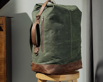 May include: A dark olive green canvas backpack featuring a brown leather base, a vertical zippered pocket on the front, a brown leather side handle, and brown leather straps. The bag has a roll-top closure and is positioned upright on a wooden stool.
