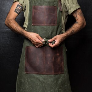 May include: A green canvas apron with two large brown leather pockets. The apron has adjustable straps and a leather buckle.