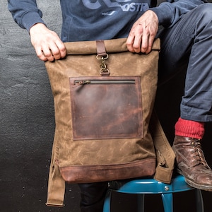 Könnte beinhalten: Ein Rolltop-Rucksack aus braunem gewachstem Canvas mit einer dunkelbraunen Leder-Fronttasche und einem horizontalen Reißverschluss. Die Tasche verfügt über einen zentralen Lederriemen mit Metallhakenverschluss sowie einen verstärkten Boden aus Leder.